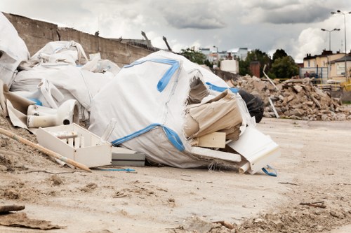 A large pile of discarded construction waste and debris piled on a dirt surface at an outdoor site, featuring white plastic sheeting, broken pipes, and various broken building materials, with a construction barrier and distant buildings visible under a cloudy sky. The scene suggests an area awaiting removal or clearance, typical of rubbish removal services in Barkingside, and showcases the type of waste handled by Commercial Waste Removal Barkingside to facilitate site cleanup and disposal. The waste is haphazardly piled, with some items partially covered by the plastic sheeting, and the ground appears dusty and unpaved, indicating an active construction or renovation environment. In the background, a wall and an urban environment with street lights and trees can be seen, emphasizing the outdoor setting within a residential or commercial district in the area. This detailed visual description reflects the type of rubbish that specialists in waste management services handle for clients in and around Barkingside.