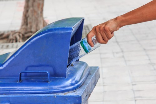 A person's hand is seen gripping a plastic water bottle and disposing of it into a blue rubbish bin with an open lid, positioned outdoors on a paved surface. The bin is made of durable plastic with a textured finish, and it appears to be part of a waste collection service offered by Commercial Waste Removal Barkingside. In the background, there is a tree trunk and a section of pavement, suggesting a public or residential area in or near Barkingside. The hand is partially visible, with only the wrist and fingers shown, and the person is wearing a short-sleeved garment. The scene captures a moment of waste disposal, highlighting proper rubbish segregation and collection in a typical urban environment within the local postcode area, relevant to rubbish removal services in Barkingside, suitable for inclusion on a commercial waste management website.