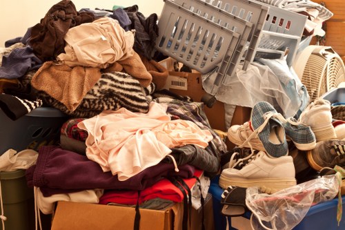 A large amount of clutter consisting of various household items and textiles piled on top of each other in an indoor or garage space. Visible are disorganized clothing and fabric items, including shirts, trousers, and towels, with some folded and others crumpled. There are also several pairs of shoes, with some in a plastic bag, casual footwear such as sneakers and sandals located at the bottom right of the pile. In the background, a grey plastic laundry basket tipped over contains more clothes, and a beige radiator is partially visible behind the clutter. The scene suggests a general rubbish or waste removal collection, typical of a professional service such as Commercial Waste Removal Barkingside, which handles bulky waste and household refuse. The environment is characterized by a chaotic arrangement, with materials and objects occupying multiple levels and corners of the space, highlighting the need for efficient rubbish clearance in the Barkingside area, within the postal code vicinity.