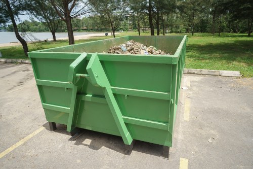 A large, open-top green skip container filled with mixed waste materials, including cardboard, plastic bags, and general rubbish, situated on a paved parking area near a park with trees and grass visible in the background. The skip has metal reinforcements on the sides and is positioned close to the curb, indicating it is prepared for rubbish removal services, such as those provided by Commercial Waste Removal Barkingside. The environment appears bright with natural daylight, highlighting the textures of the waste and the smooth, painted surface of the skip, which contrasts with the surrounding greenery and paved surroundings.
