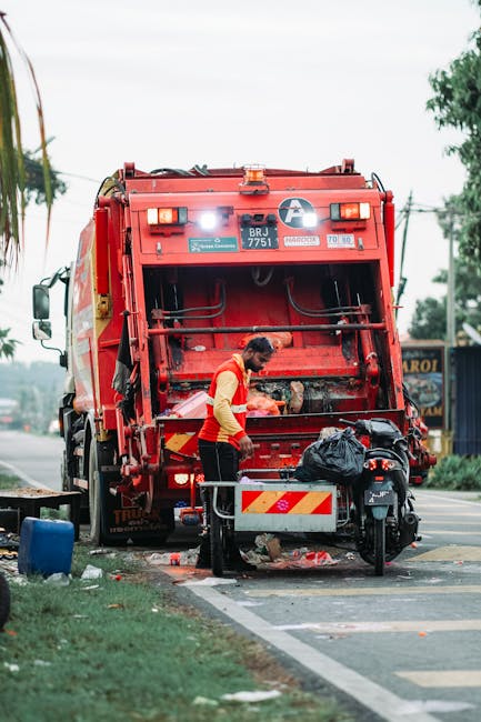 A waste collection worker is standing behind a large red garbage truck on a roadside, meticulously emptying a black rubbish bag into the truck’s open rear compartment. The worker is wearing a high-vis