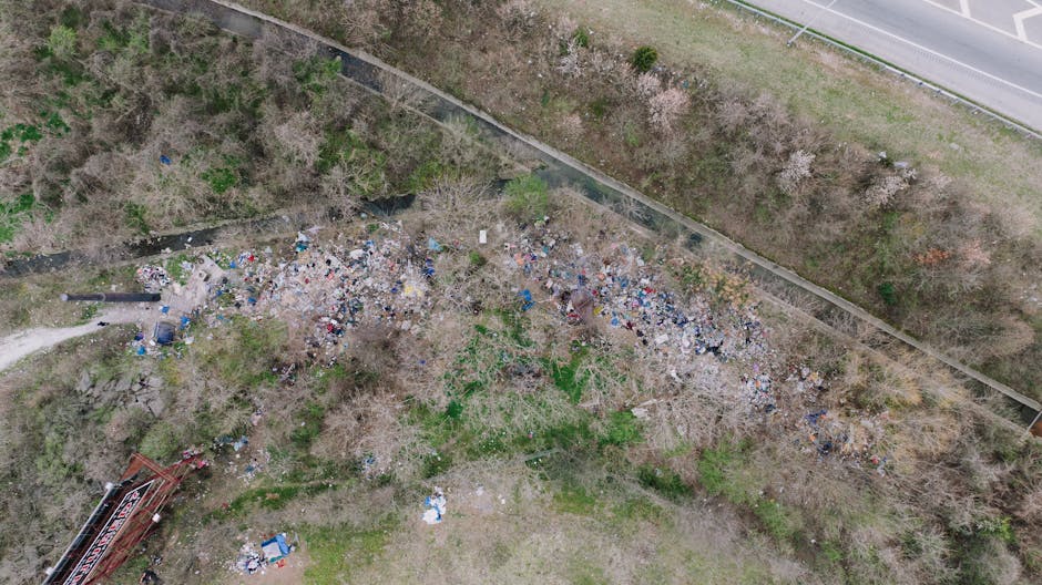 An aerial view of a large, disorganized accumulation of mixed waste and debris scattered across a patch of bare ground next to a narrow pathway, with sparse trees and shrubs surrounding the area. The 