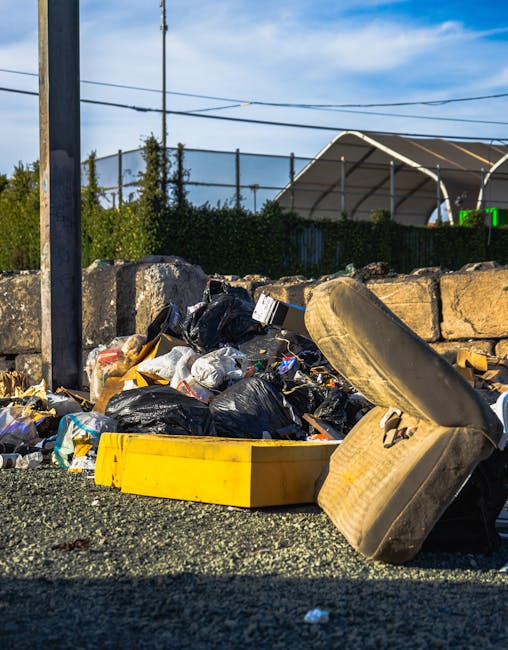 An aerial view of a large, disorganized accumulation of mixed waste and debris scattered across a patch of bare ground next to a narrow pathway, with sparse trees and shrubs surrounding the area. The 